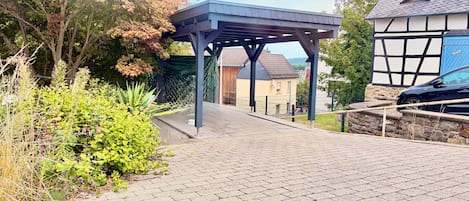 En asfalteret sti der fører hen til en overdækket bygning, med udsigt til boliger og en parkeret bil i nærheden.