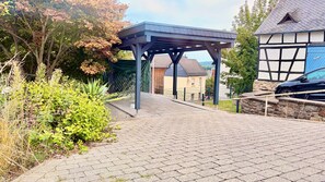A paved pathway leading to a covered structure with a view of residential buildings and a car parked nearby.