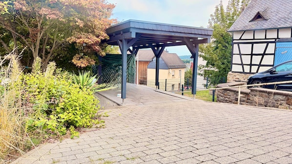 A paved pathway leading to a covered structure with a view of residential buildings and a car parked nearby.