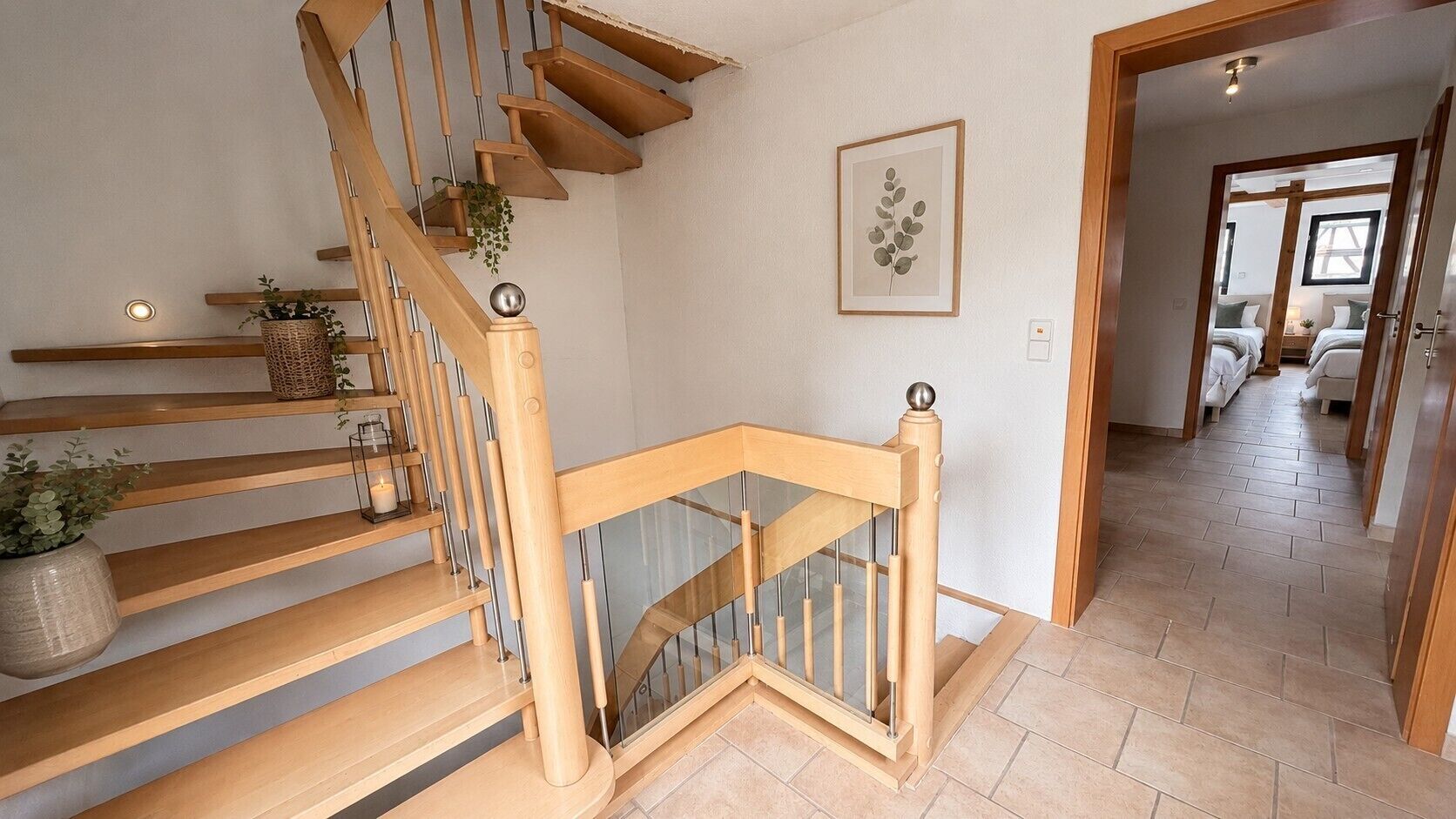 A wooden staircase with glass railings, a framed botanical print on the wall, and a hallway leading to a bedroom.