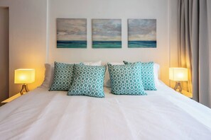 Een hotelkamer met een bed, vier gestreepte kussens, twee nachtlampen en drie aan de muur hangende schilderijen van een zeegezicht.