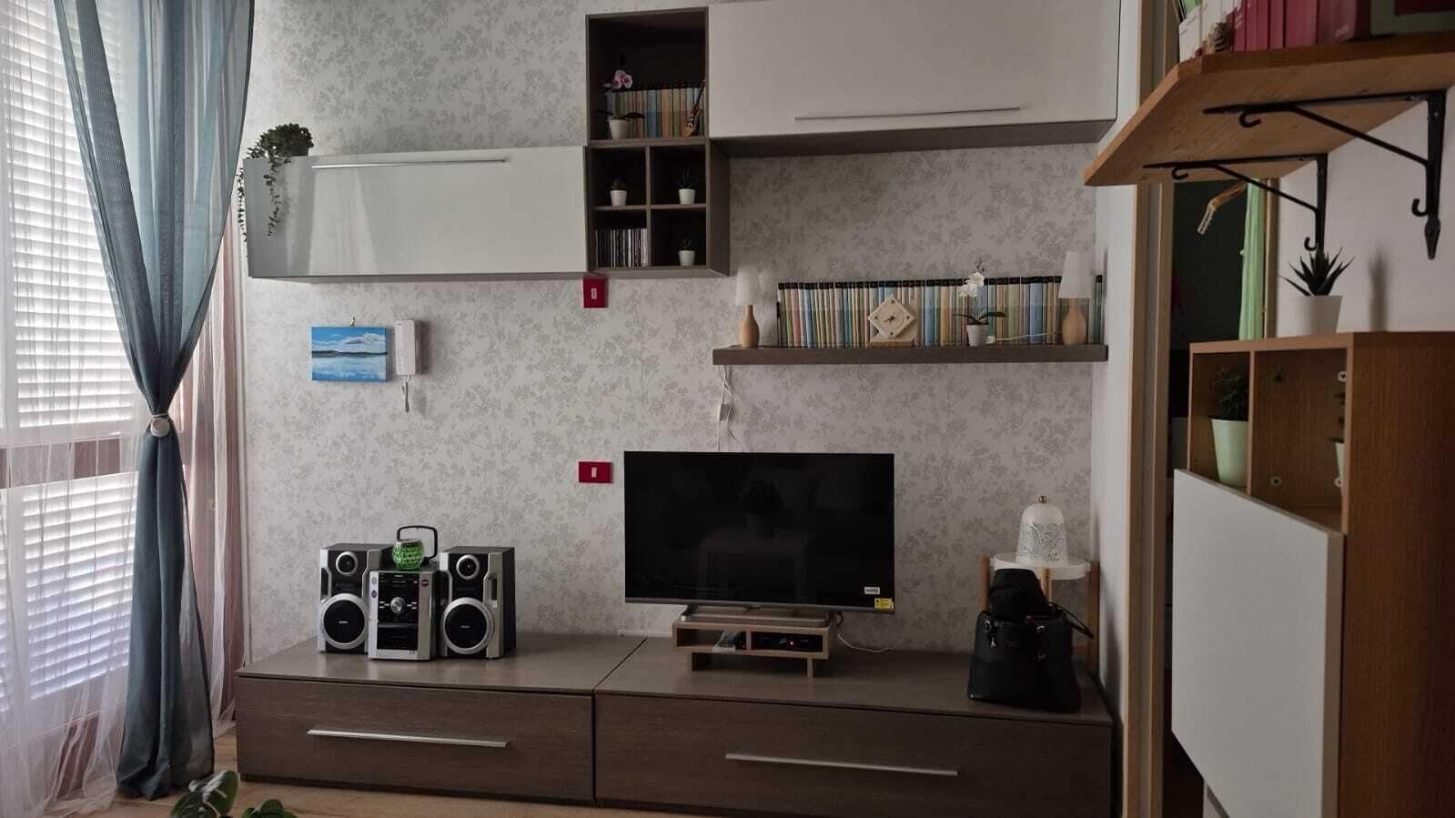 Ein Wohnzimmer mit Flachbildfernseher, zwei Lautsprechern, einer Bücherwand und einem kleinen Tisch mit einer Lampe.