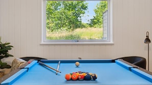 Ein Billardtisch mit Kugeln, einem Queue und einem Fenster mit Blick auf Bäume.