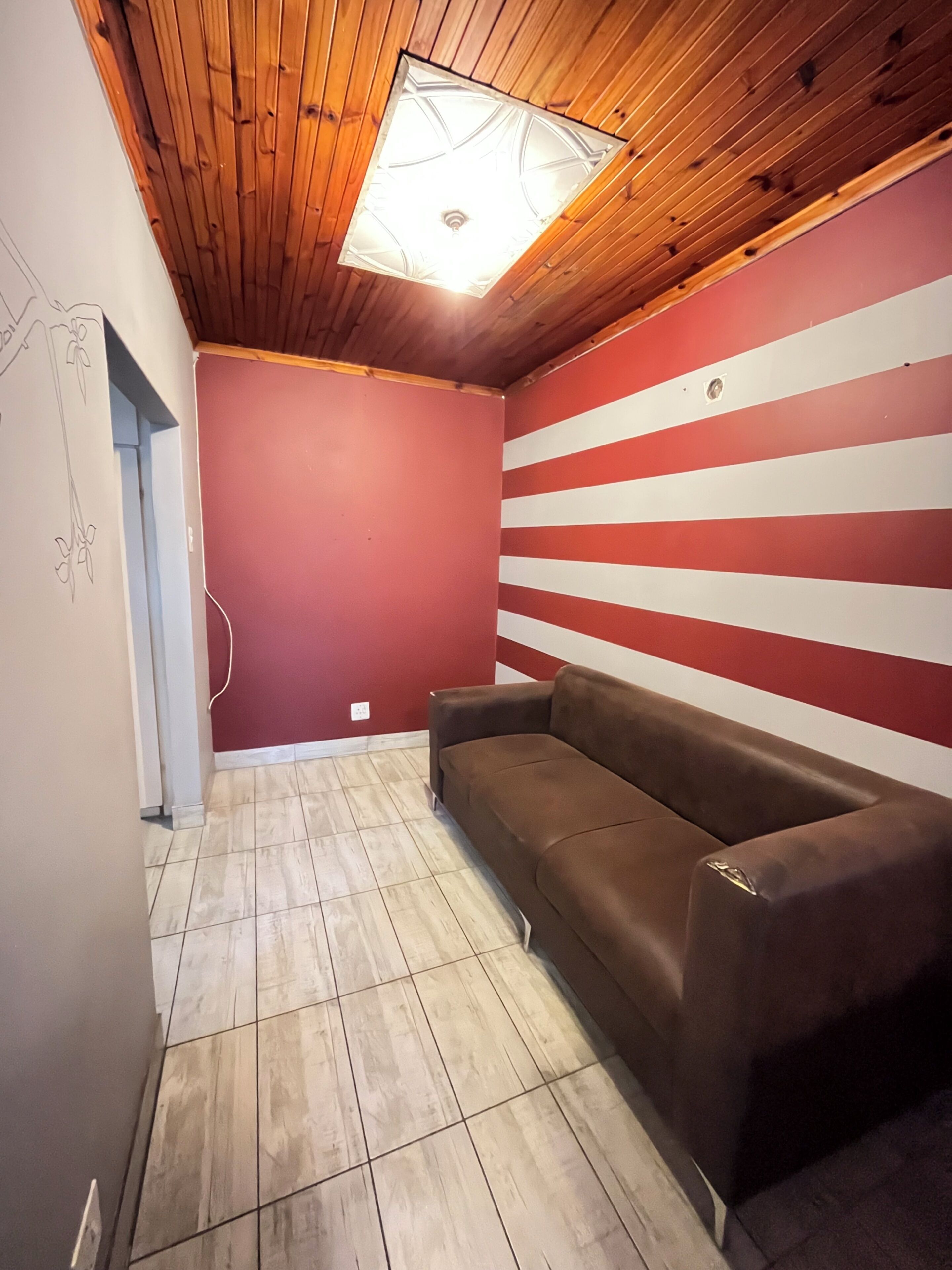 Une pièce avec un plafond en bois, un mur à rayures rouges et blanches, un canapé marron et un sol carrelé.