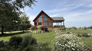 Une maison en bois dotée d’un grand porche et d’un espace couvert, entourée d’un jardin bien entretenu où poussent différentes espèces de plantes et où s’étend une pelouse.