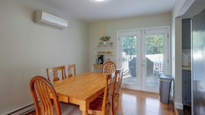 Een eetgedeelte met een houten tafel en stoelen, een airconditioner aan de muur, een koffiezetapparaat op een plank en uitzicht door een glazen deur.