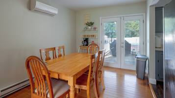 Un coin repas avec une table et des chaises en bois, un climatiseur fixé au mur, une machine à café posée sur une étagère et une vue à travers une porte vitrée.