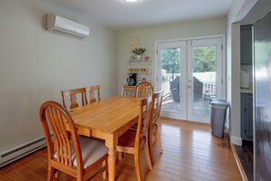 Een eetgedeelte met een houten tafel en stoelen, een airconditioner aan de muur, een koffiezetapparaat op een plank en uitzicht door een glazen deur.