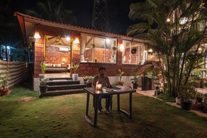 Een bakstenen huis met een veranda, plantenbakken en een tafel waarop iemand zit.