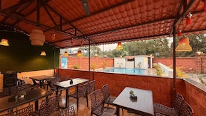 An outdoor seating area with tables and chairs, a pool in the background, and a covered roof structure.