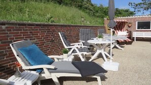 Eine Terrasse mit weißen Liegestühlen und einem Tisch, eine Ziegelwand und ein kleines Gebäude im Hintergrund.
