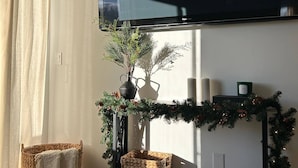 A living room with a flat-screen TV mounted on the wall, a Christmas garland, two wicker baskets, and a large window with curtains.