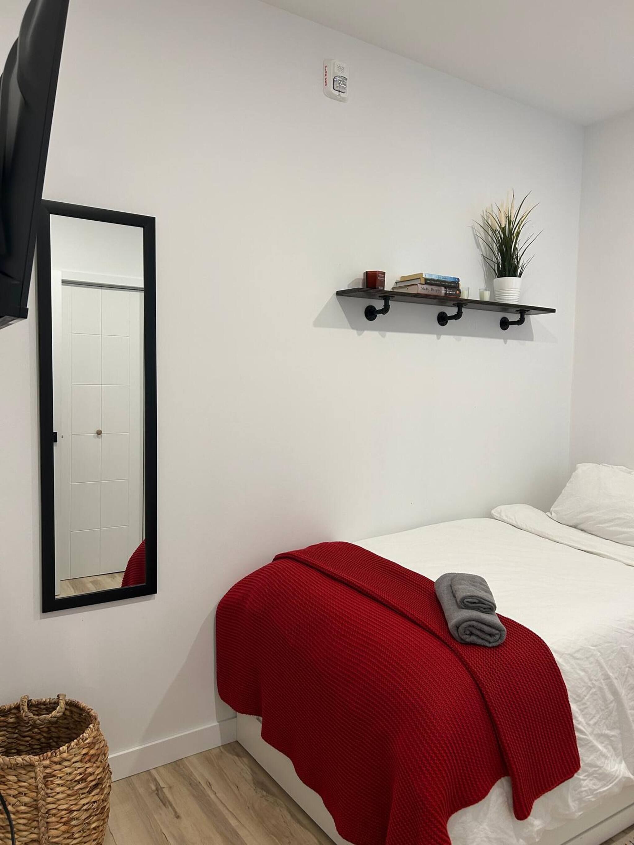 Une chambre à coucher avec un lit, une couverture rouge, un miroir, une étagère avec des livres et une plante, et un panier.