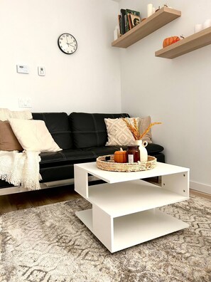 A modern living room with a black leather sofa, a white coffee table with a woven basket, a wall clock, and floating shelves with books and pumpkins.