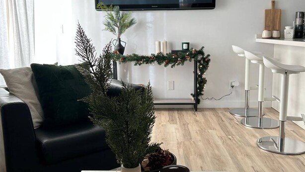 Ein modernes Wohnzimmer mit einem an der Wand befestigten Flachbildfernseher, einer schwarzen Ledersofa, einem weißen Couchtisch mit Dekoration und einem Essbereich mit weißen Barhocker.