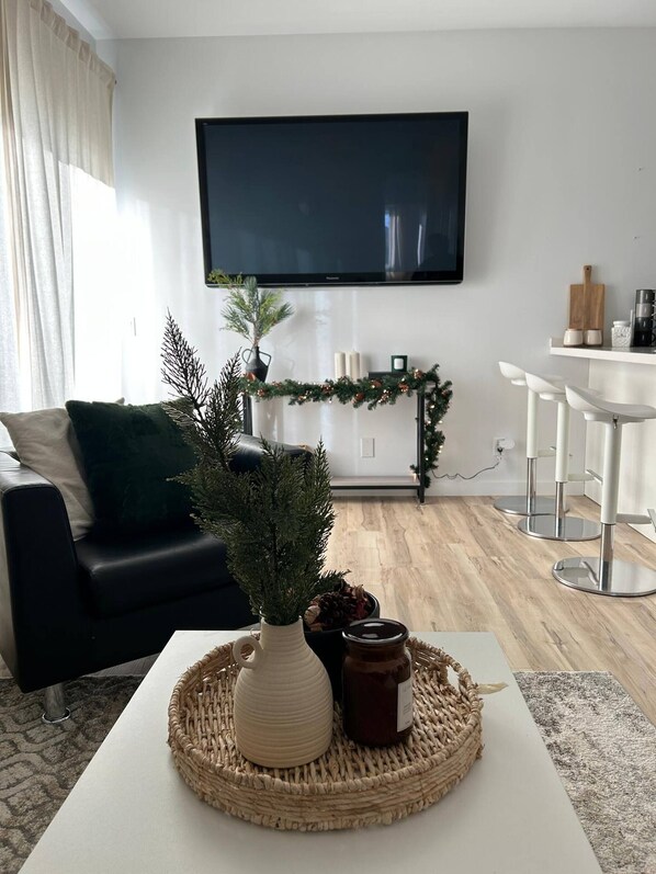 A modern living room with a flat-screen TV mounted on the wall, a black leather sofa, a white coffee table with decorative items, and a dining area with white bar stools.