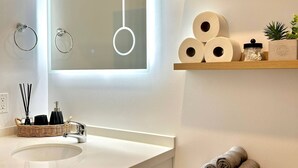 Ein Badezimmer mit weißem Waschbecken, einem Spiegel mit Beleuchtung, einer Toilette und Handtüchern auf einem Regal.
