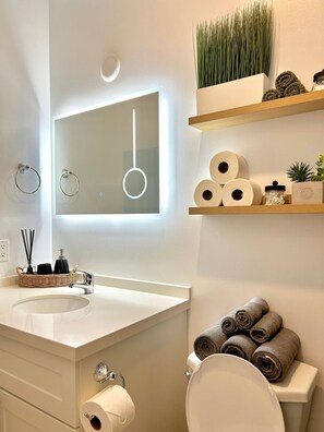 A bathroom with a white sink, a mirror with a light, a toilet, and towels on a shelf.