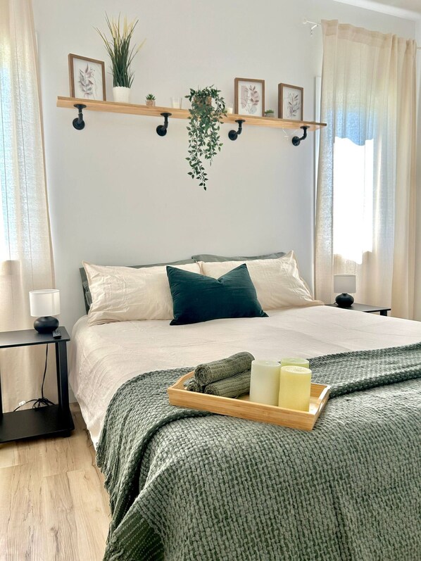 A neatly arranged bedroom with a bed, bedside tables, a tray with towels and candles, and a wall-mounted shelf with plants and pictures.