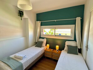 Een tweepersoonskamer met twee bedden, een raam met gordijnen, bedlampjes en een kleine plant aan de muur.