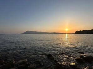 Rustig water met een ondergaande zon op de achtergrond en een rotsachtige kustlijn op de voorgrond.