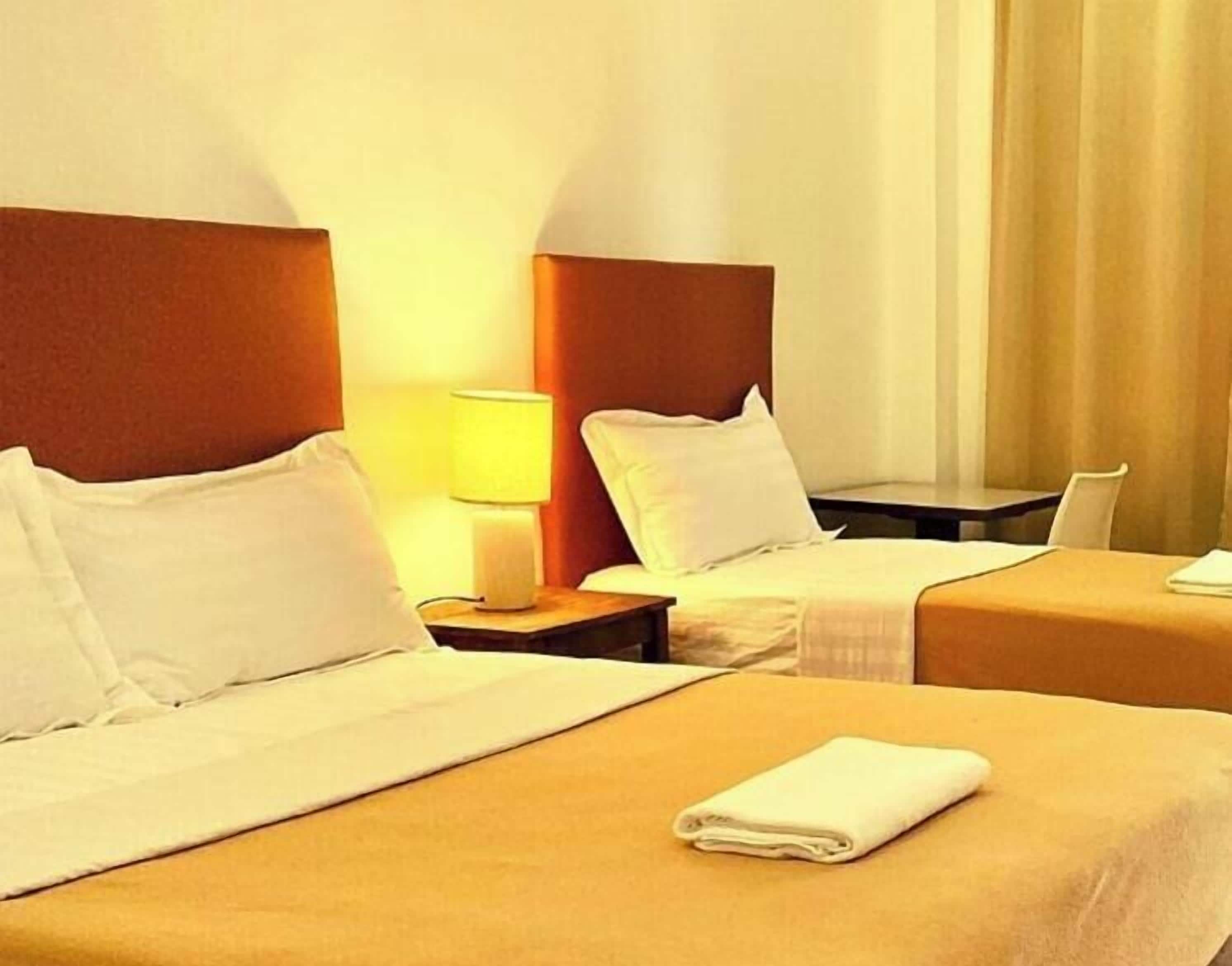 A hotel room with two beds, a nightstand with a lamp, and a view of the outside.