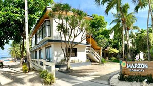 A modern two-story building with a white facade and large windows, surrounded by palm trees and a beach view.
