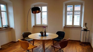 Quarto com mesa de madeira redonda, quatro cadeiras, luminária de chão e duas janelas com vista para edifícios.