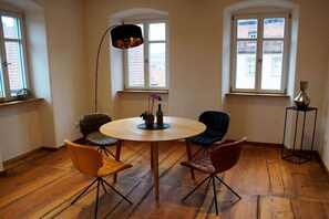 Ett rum med ett runt träbord, fyra stolar, en golvlampa och två fönster med utsikt över byggnader.