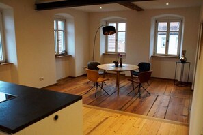 Un coin repas comprenant une table ronde et quatre chaises, un plancher en bois et deux fenêtres équipées de volets.