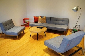 Een woonkamer met een grijze bank, twee grijze fauteuils, een ronde houten salontafel en een rode bijzettafel.