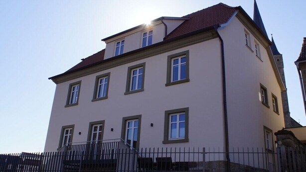 Ein mehrgeschossiges Gebäude mit einer symmetrischen Fassade, einem roten Ziegeldach und einem freistehenden Kirchturm.
