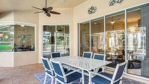 Un espace patio aménagé avec une table et des chaises, un ventilateur de plafond et une vue sur une piscine, visible à travers de grandes portes vitrées.