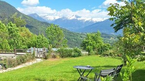 Ein grünes Freigelände mit Tisch und Stühlen, umgeben von Bergen und blauem Himmel.