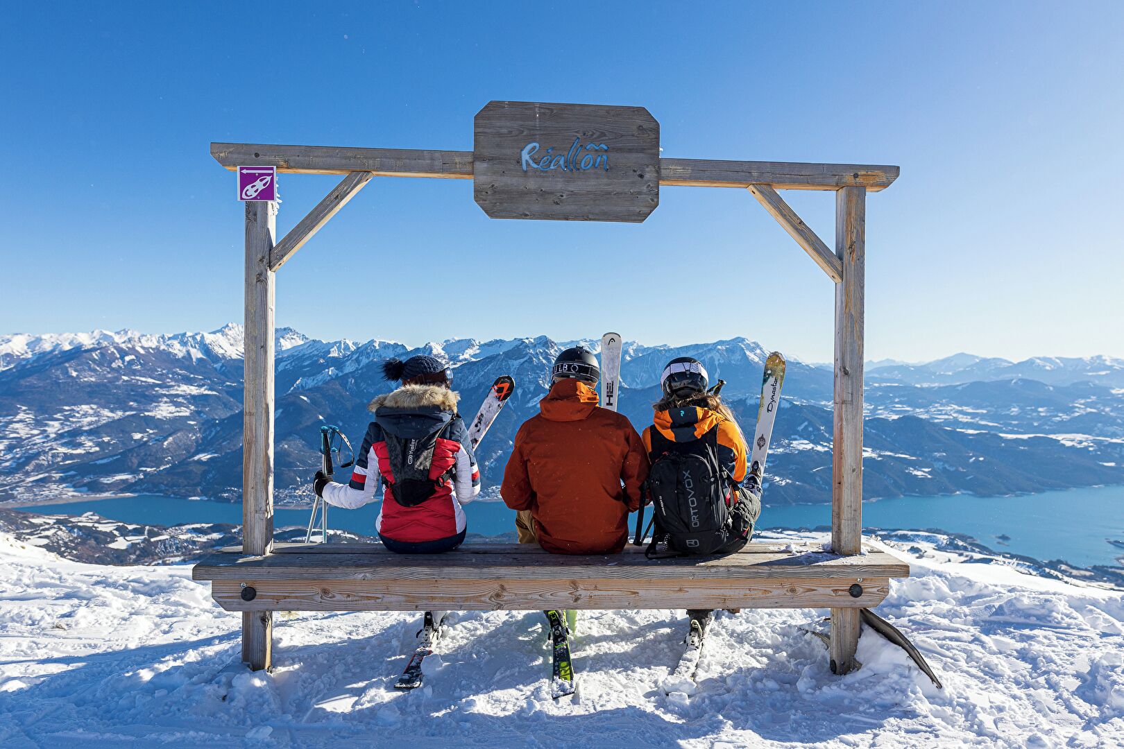 A wooden bench with a sign reading 'Réallon' in a snowy landscape with mountains and a lake in the background.