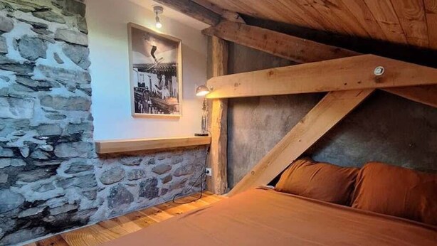 Ein Schlafzimmer mit schrägen Wänden, einer Steinwand, Holzbalken, einem Bett mit orangefarbener Bettwäsche und einem gerahmten Bild an der Wand.