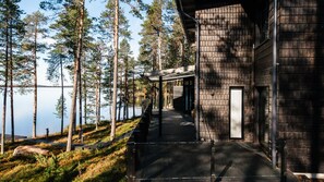 A building with a covered walkway leading to a door, surrounded by tall trees and a body of water. - Willa Lumia - Aurora Skylight Villa (Rovaniemi)