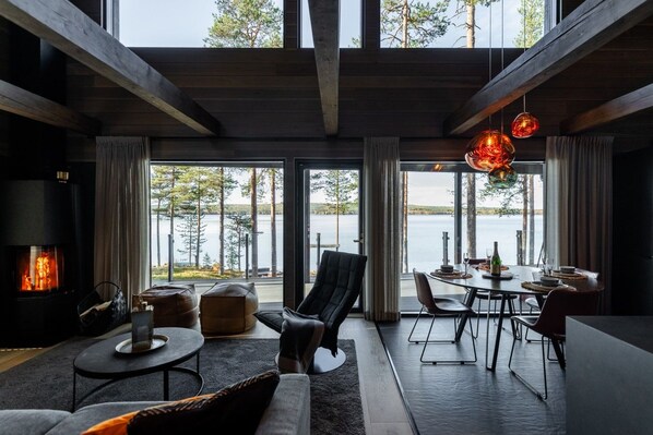 Un intérieur moderne avec une cheminée, un coin salon, une table à manger et de grandes fenêtres donnant sur un plan d’eau.