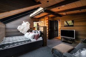 A cozy bedroom with a sloped ceiling, a bed with multiple pillows, a television, and a small seating area. - Willa Lumia - Aurora Skylight Villa (Rovaniemi)