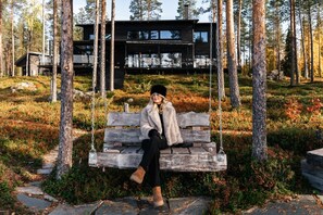A person sitting on a wooden swing in a forested area with a modern building in the background.