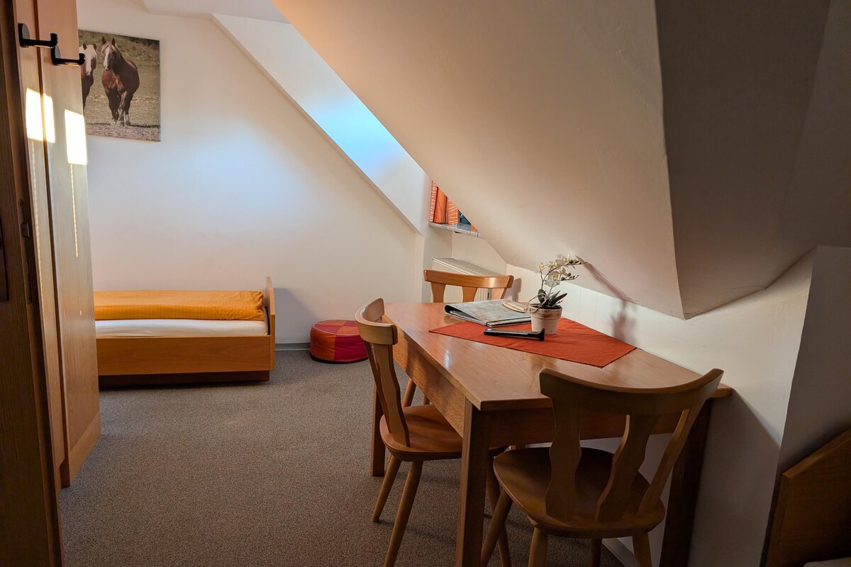 A room with a wooden bed, a dining table with chairs, a small red stool, and a potted plant.