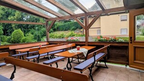 An outdoor covered patio with wooden benches and tables, surrounded by greenery and a view of a building.