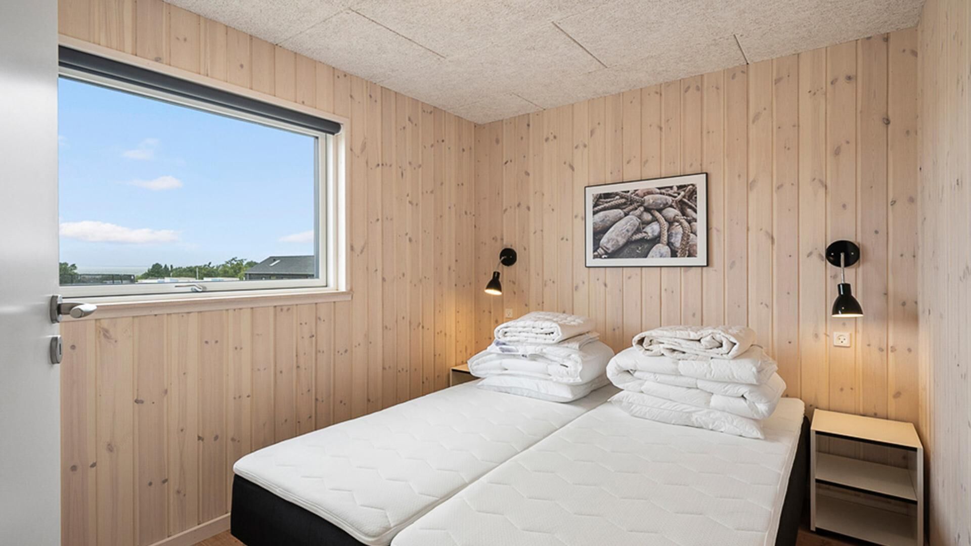 A room with wooden walls, a window, a bed with white bedding, a small bedside table, and a wall-mounted picture.