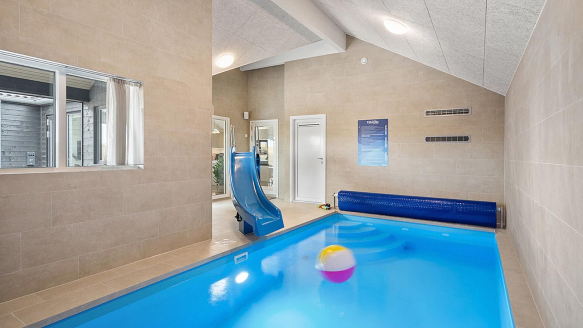 An indoor swimming pool with a slide, a poolside seating area, and a door leading to another room.