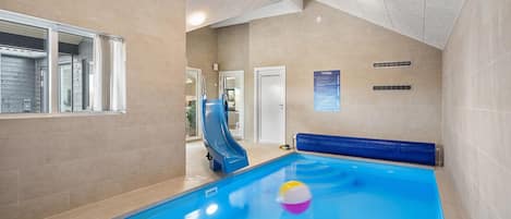 En inomhuspool med en vattenrutschkana, ett sittområde vid poolkanten och en dörr som leder till ett annat rum.