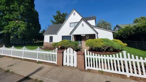 A single-story house with a white exterior, a brick foundation, and a white picket fence. - Carlton Cottage (Burney)