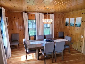 Un coin repas avec une table en bois, six chaises, un lustre et une vue sur l’extérieur grâce à de grandes fenêtres.