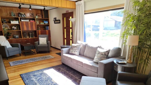 A living room with a sofa, armchairs, a coffee table, a bookshelf, and a large window with a view. - Carlton Cottage (Burney)