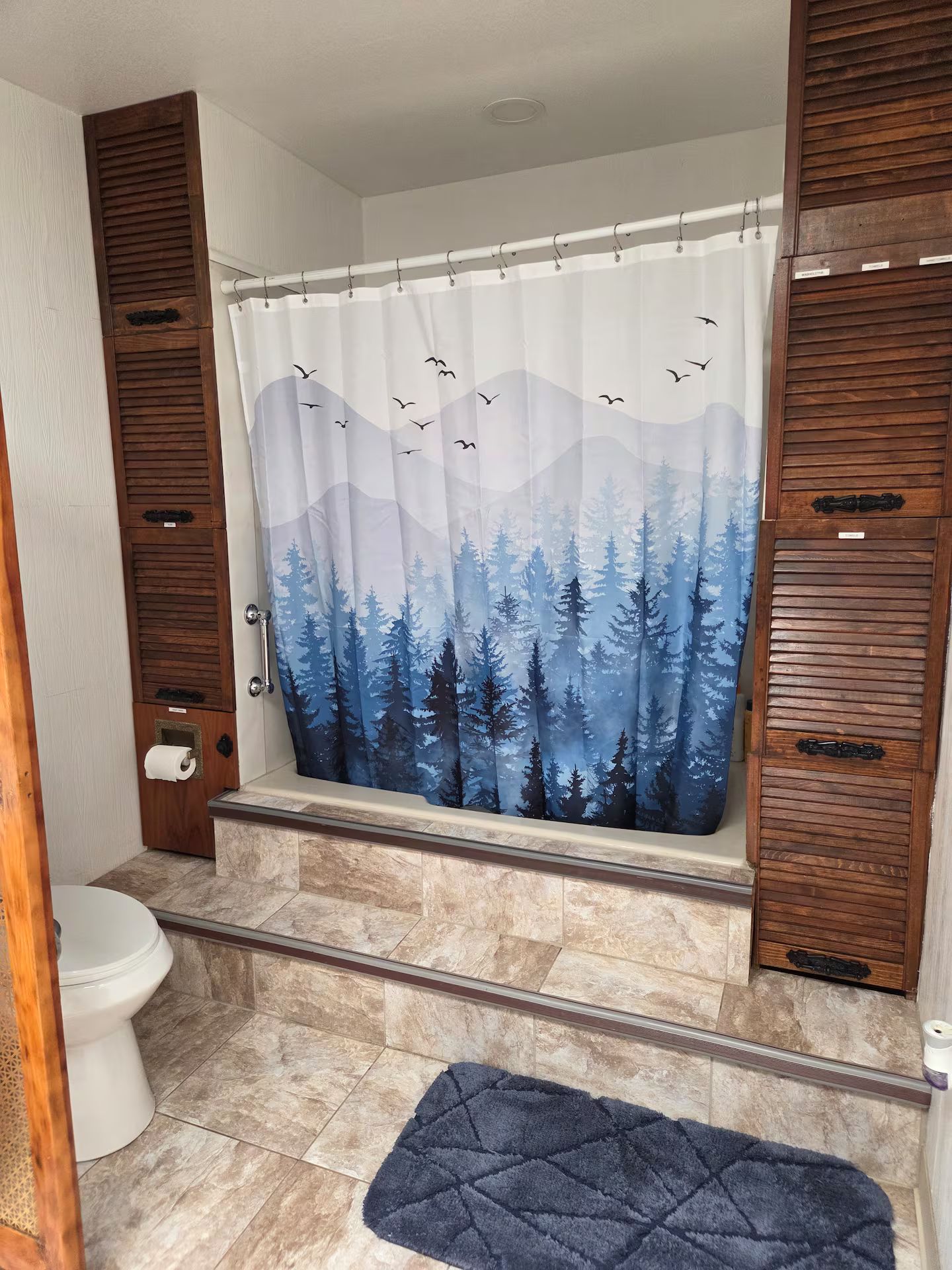 Une salle de bain avec un rideau de douche représentant une scène de forêt, des toilettes et un tapis bleu.
