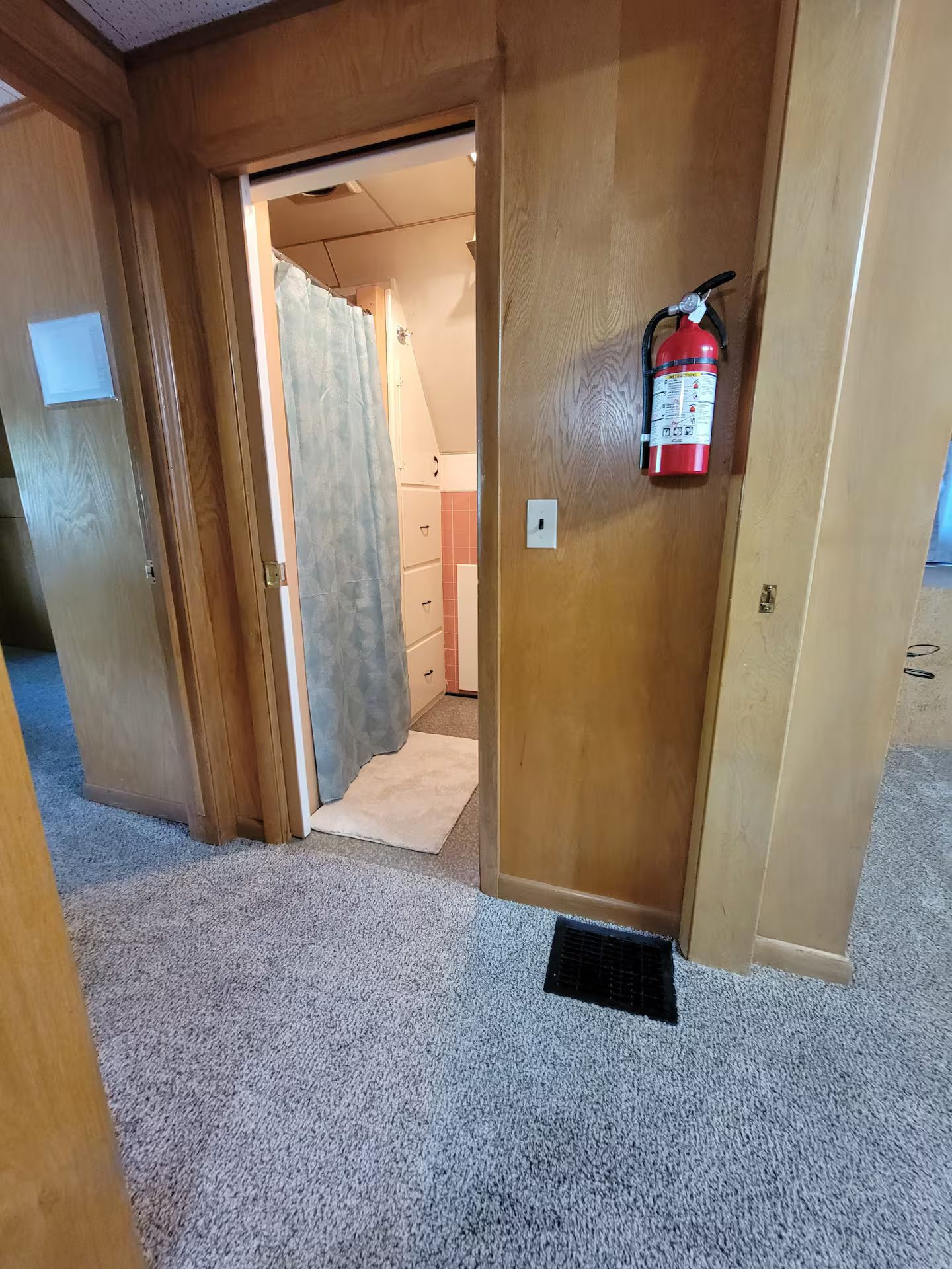 Ein Flur mit einer Holztür, einem an der Wand befestigten Feuerlöscher und einem Teppichboden.